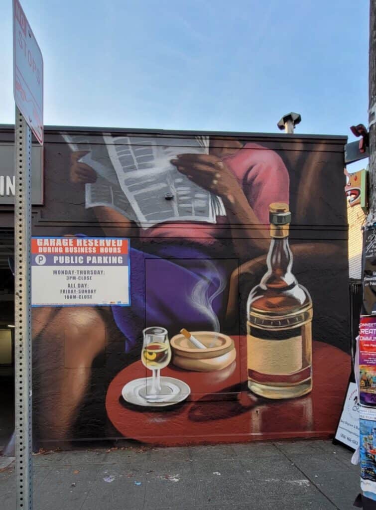 Mural on a building wall showing a person reading a newspaper at a table with a bottle of liquor, a glass, and an ashtray, next to a public parking sign.