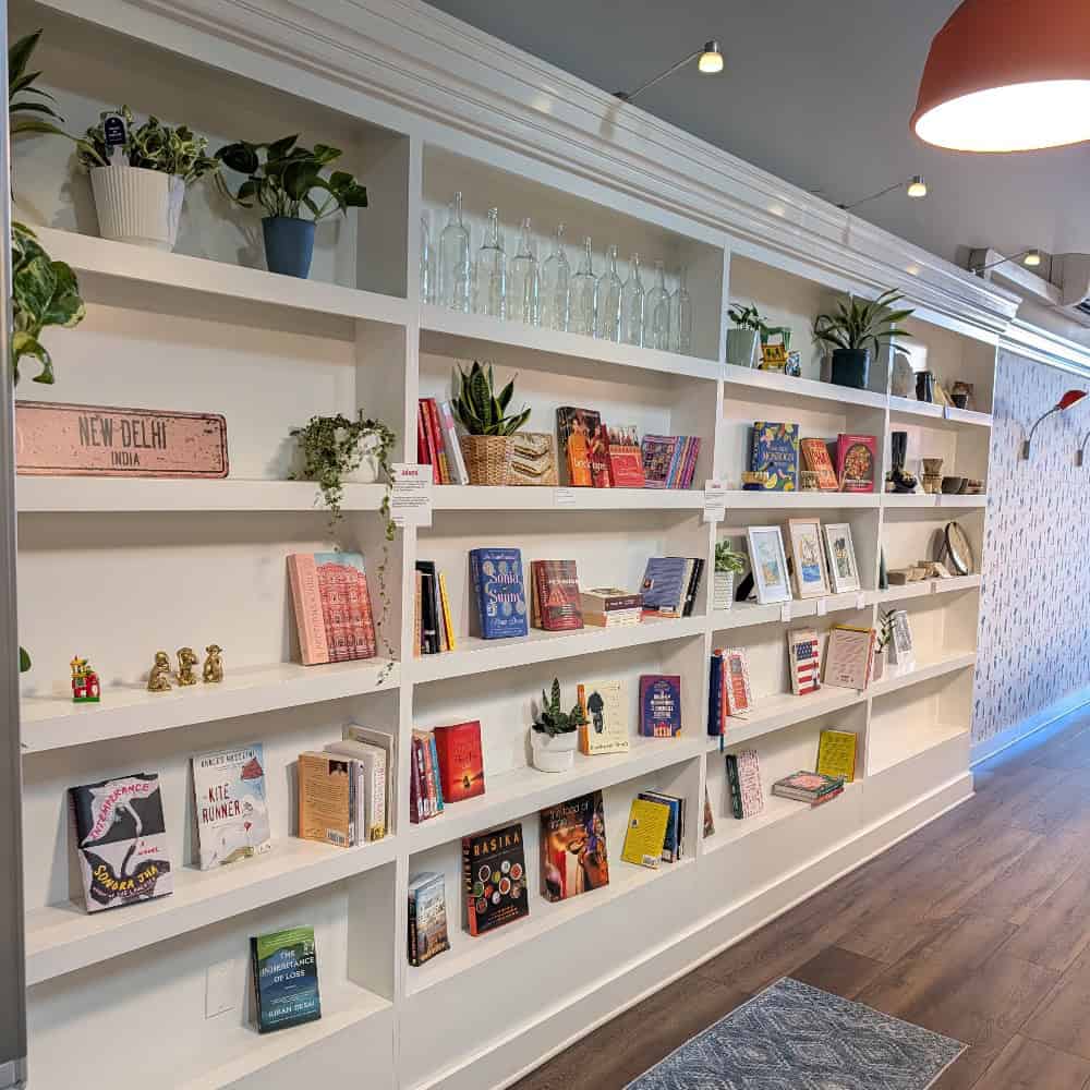 Large white built-in bookshelves at Janta cafe filled with books, potted plants, glass bottles, and a New Delhi street sign.