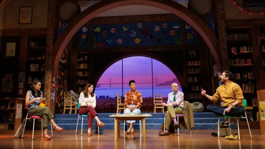Five people sit in chairs on a stage decorated as a library, with a large window backdrop showing the Golden Gate Bridge at sunset.