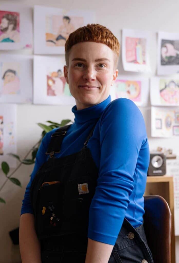 Person with short red hair wearing a bright blue turtleneck and black overalls, sitting indoors in front of a wall decorated with colorful drawings.