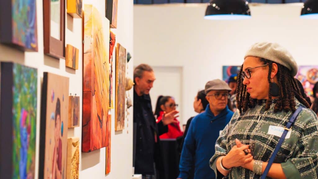 A woman wearing a beret and glasses looks at paintings displayed on a gallery wall while other people stand in the background.