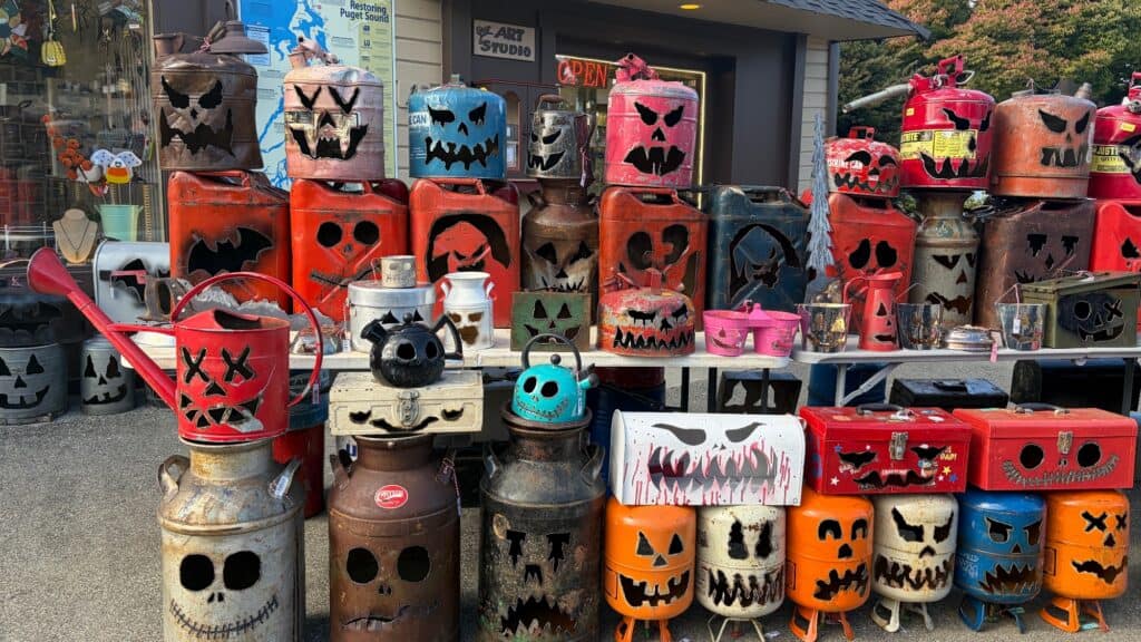 A display of repurposed metal containers, such as gas cans and canisters, carved into various jack-o’-lantern faces and placed on tables outside a shop.