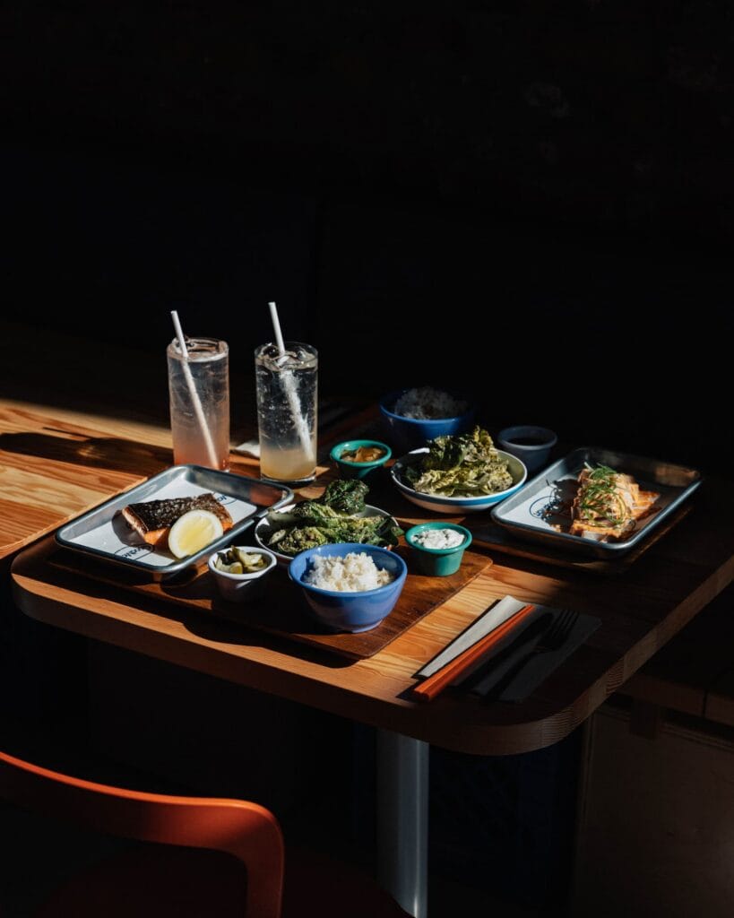 A table spread featuring two salmon fillets, bowls of rice, roasted broccoli, a small salad, and two glasses of iced drinks.