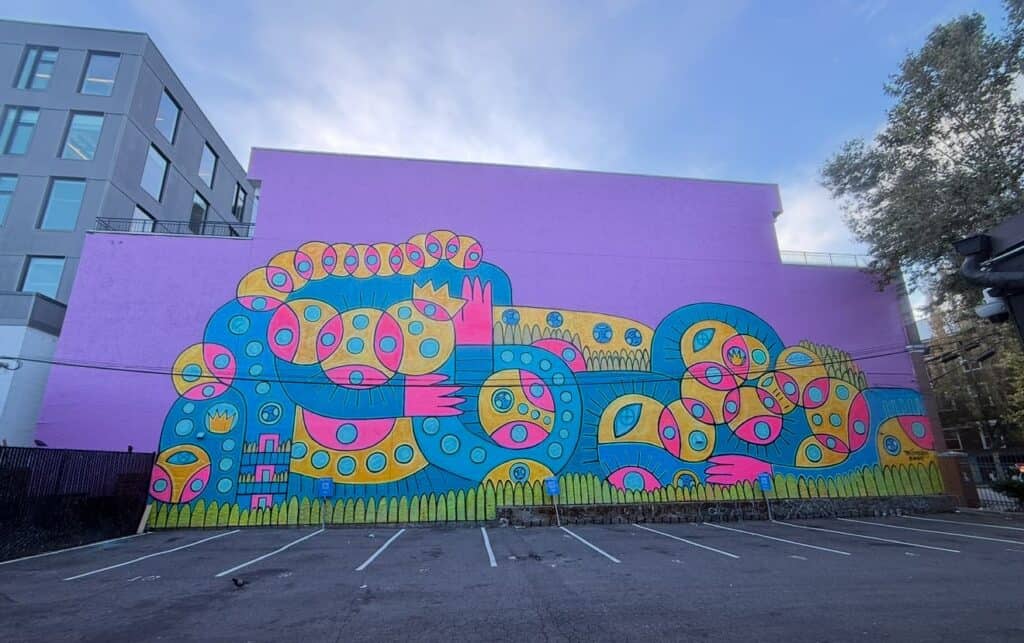 A colorful mural featuring abstract, cartoon-like figures with circular motifs painted on a large purple wall above a parking lot.