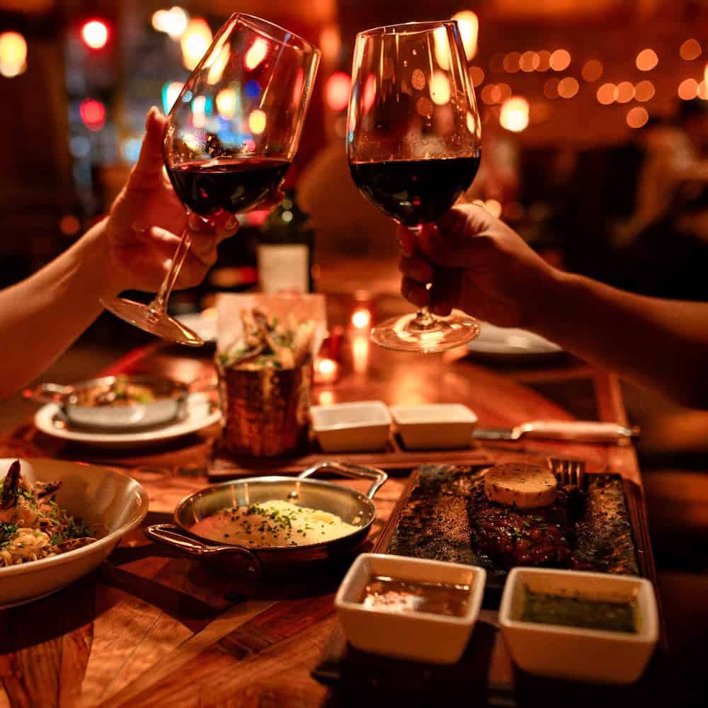 Two people clinking glasses of red wine over a restaurant table spread with steak, side dishes, and appetizers.