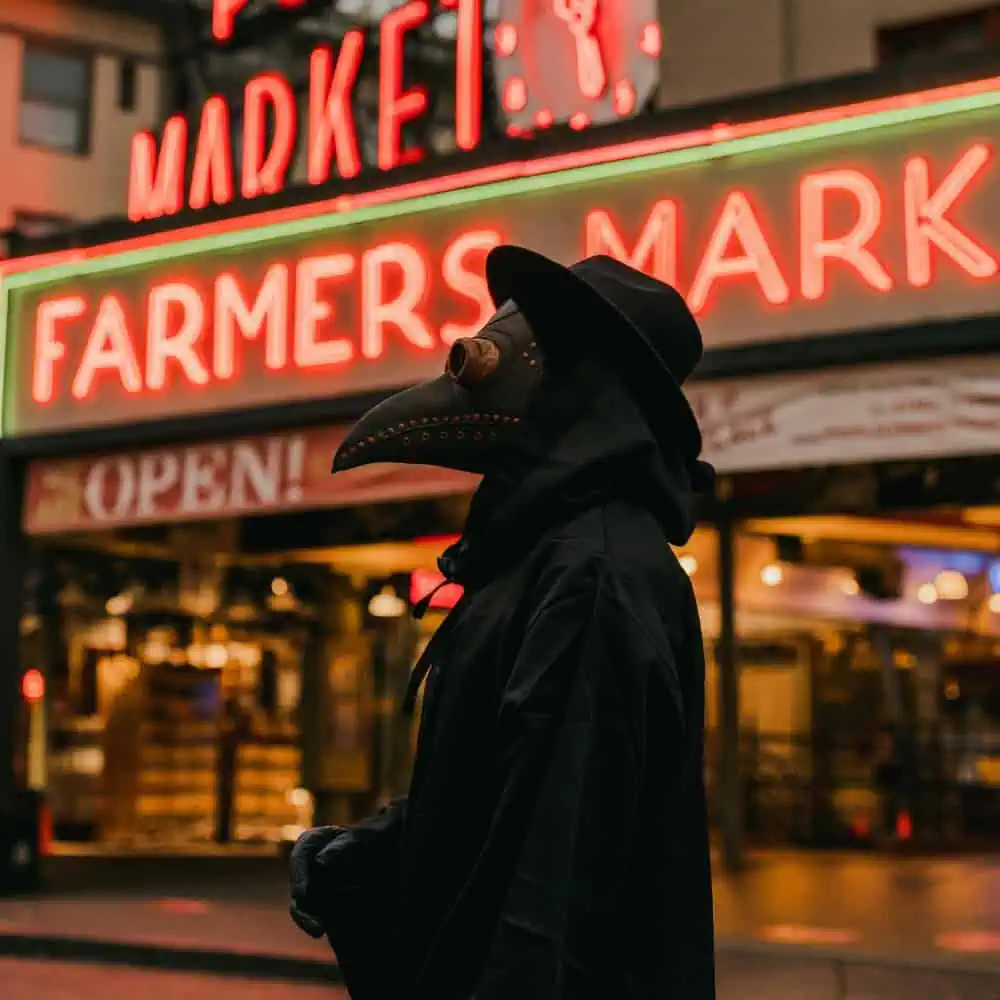 A person in a plague doctor mask and hooded cloak standing in front of a neon “Farmers Market” sign at night.