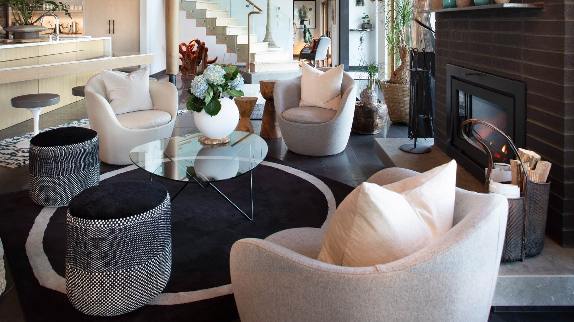 Modern living room with stylish furniture, including plush chairs, a glass coffee table, and a fireplace, featuring elegant decor and an open kitchen in the background.