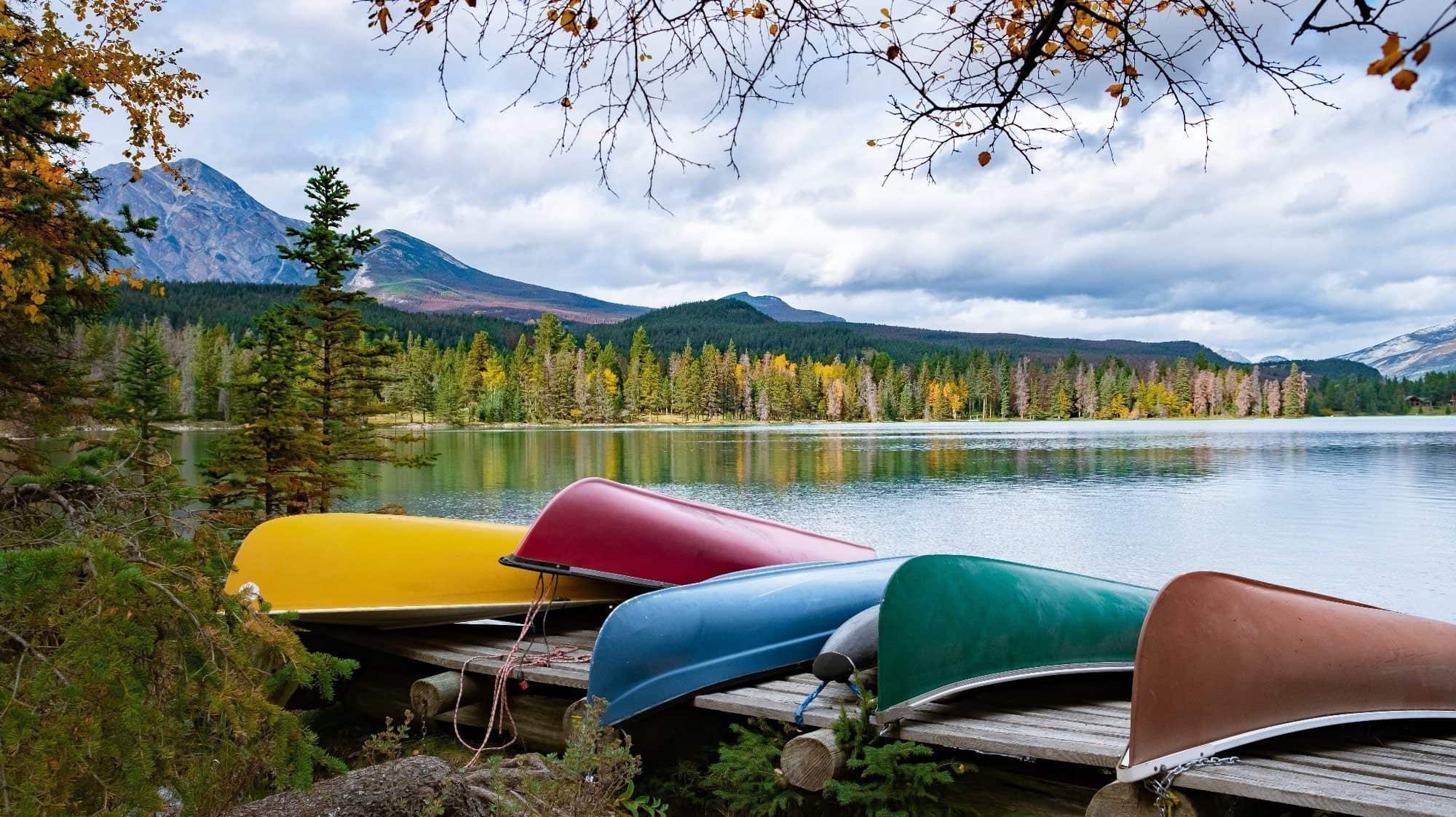 Colorful canoes stacked on a wooden dock beside a serene lake, with autumn trees and mountains in the background, creating an atmosphere of peace.