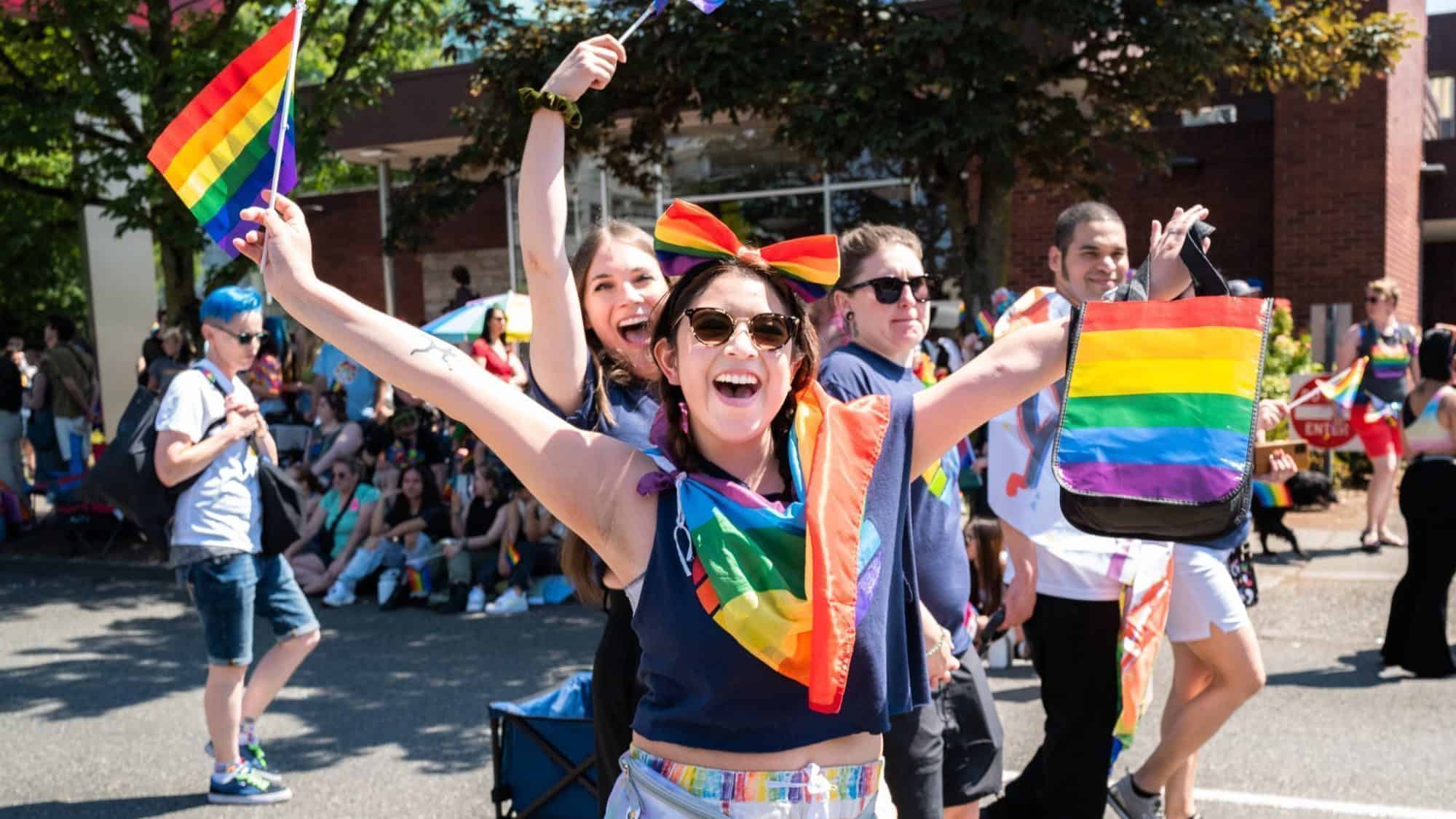 People are celebrating at an outdoor event, holding rainbow flags and wearing colorful attire. The festive scene, marking 50 Years of Seattle Pride, is filled with smiles against a backdrop of trees.