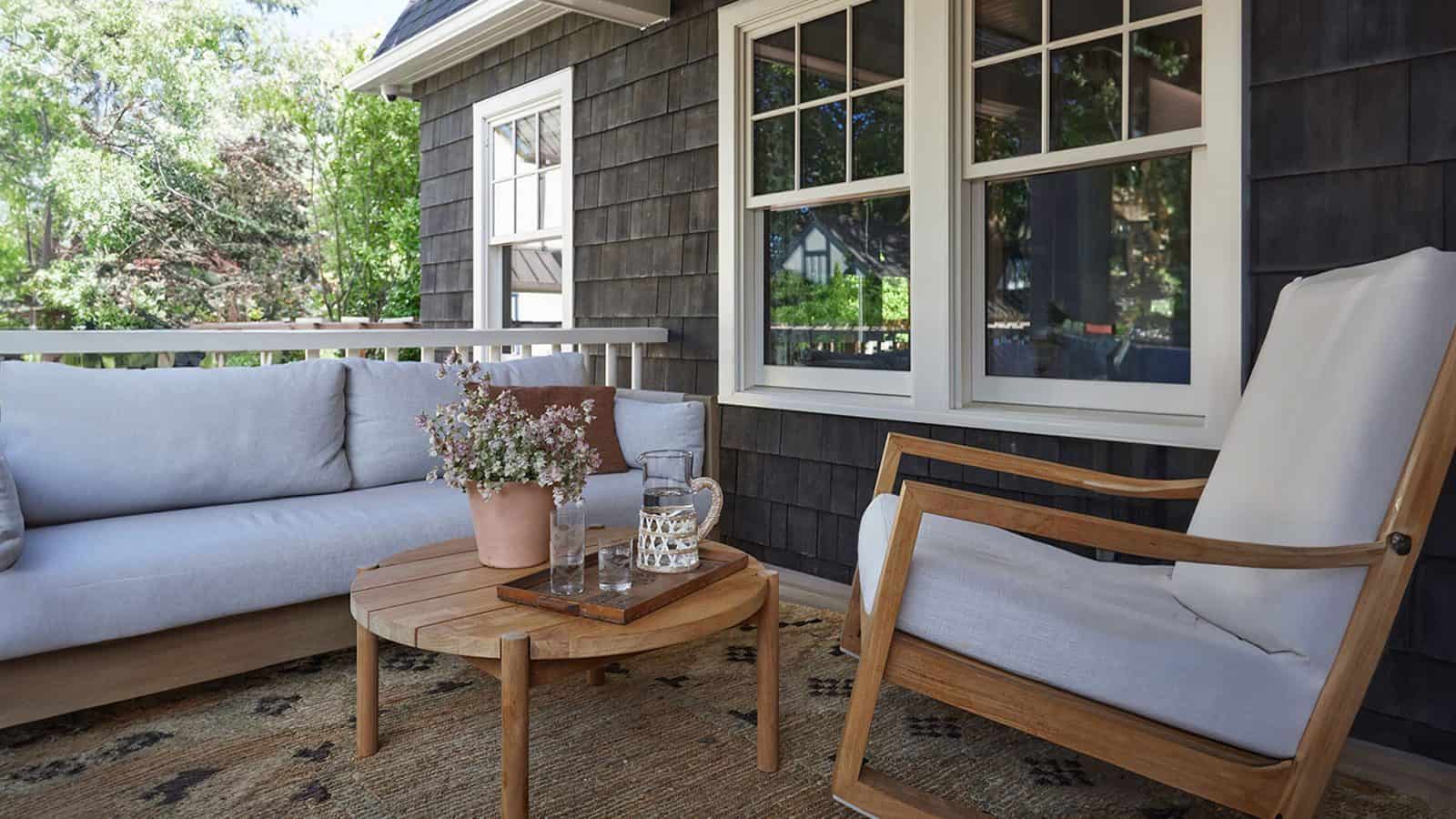 An outdoor patio for maximalists features a cushioned sofa, a wooden rocking chair, and a round wooden coffee table with a potted plant and glassware, all under the serene Montlake sky.