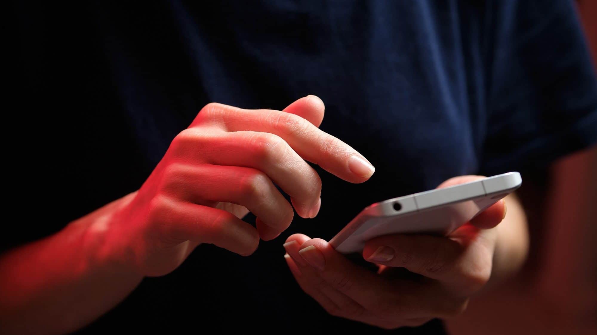 A person in a dark shirt is holding and using a smartphone with both hands, illuminated by red lighting, showcasing the bad habit of always checking your phone.