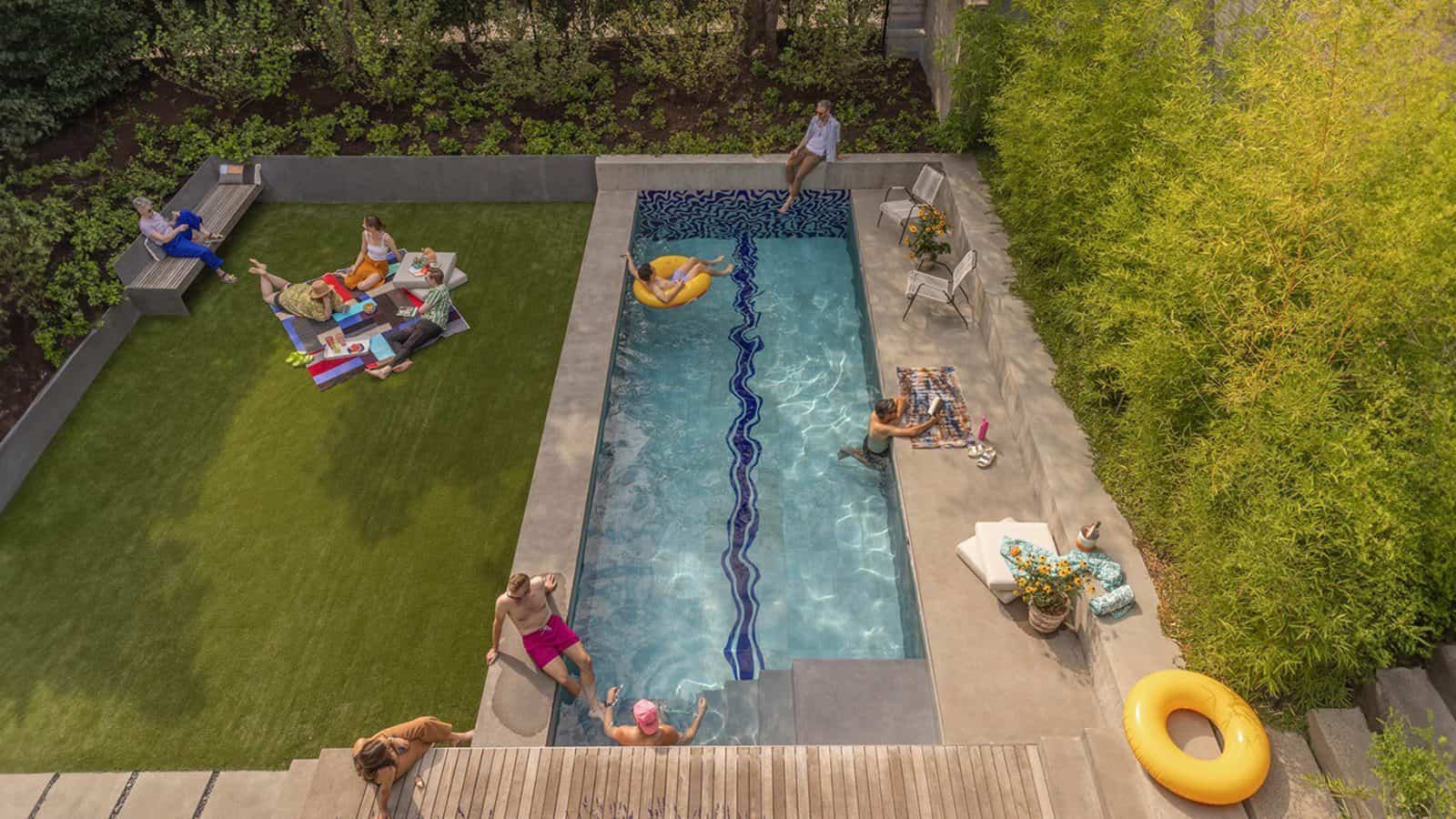 The Leschi backyard features a rectangular pool with people swimming and lounging on inflatables, surrounded by greenery. Others sit on the grass and by the patio, enjoying the sunny day, creating an immersive experience for everyone.