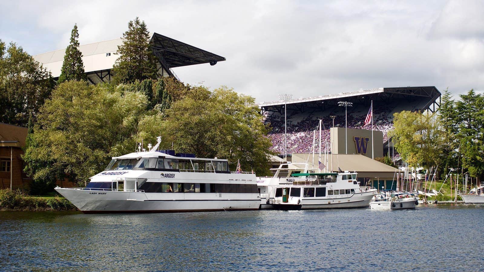 Boats docked on a river with trees and a stadium in the background, creating the perfect setting for sailgating enthusiasts.