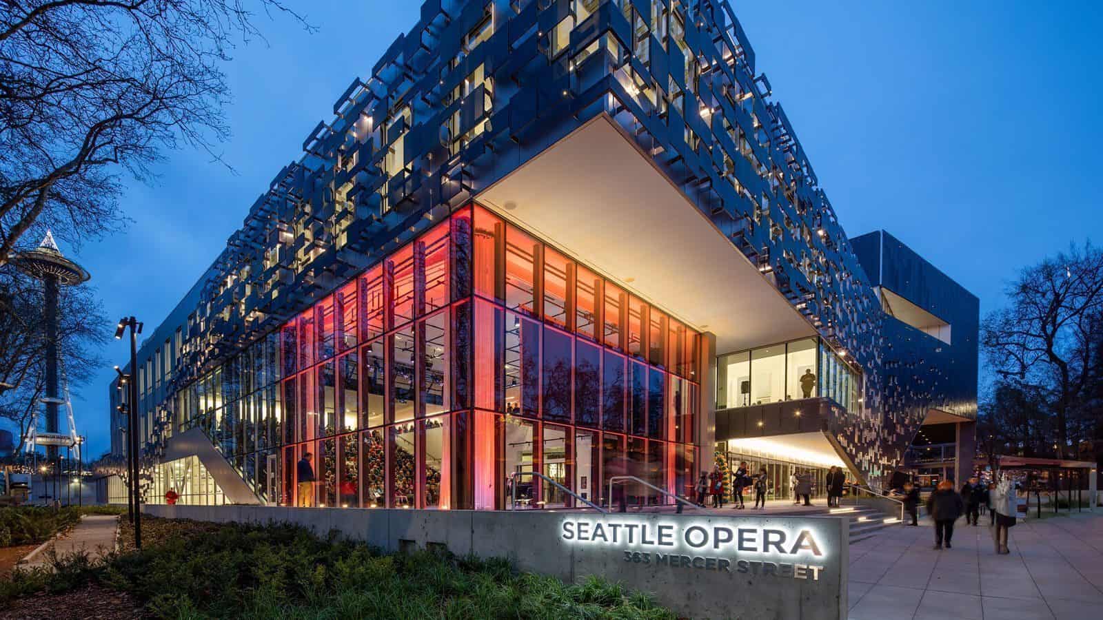 Exterior view of the Seattle Opera building at 363 Mercer Street, illuminated at dusk. The modern architecture, worthy of a maestro's appreciation, features large glass windows and a unique façade with patterned panels.