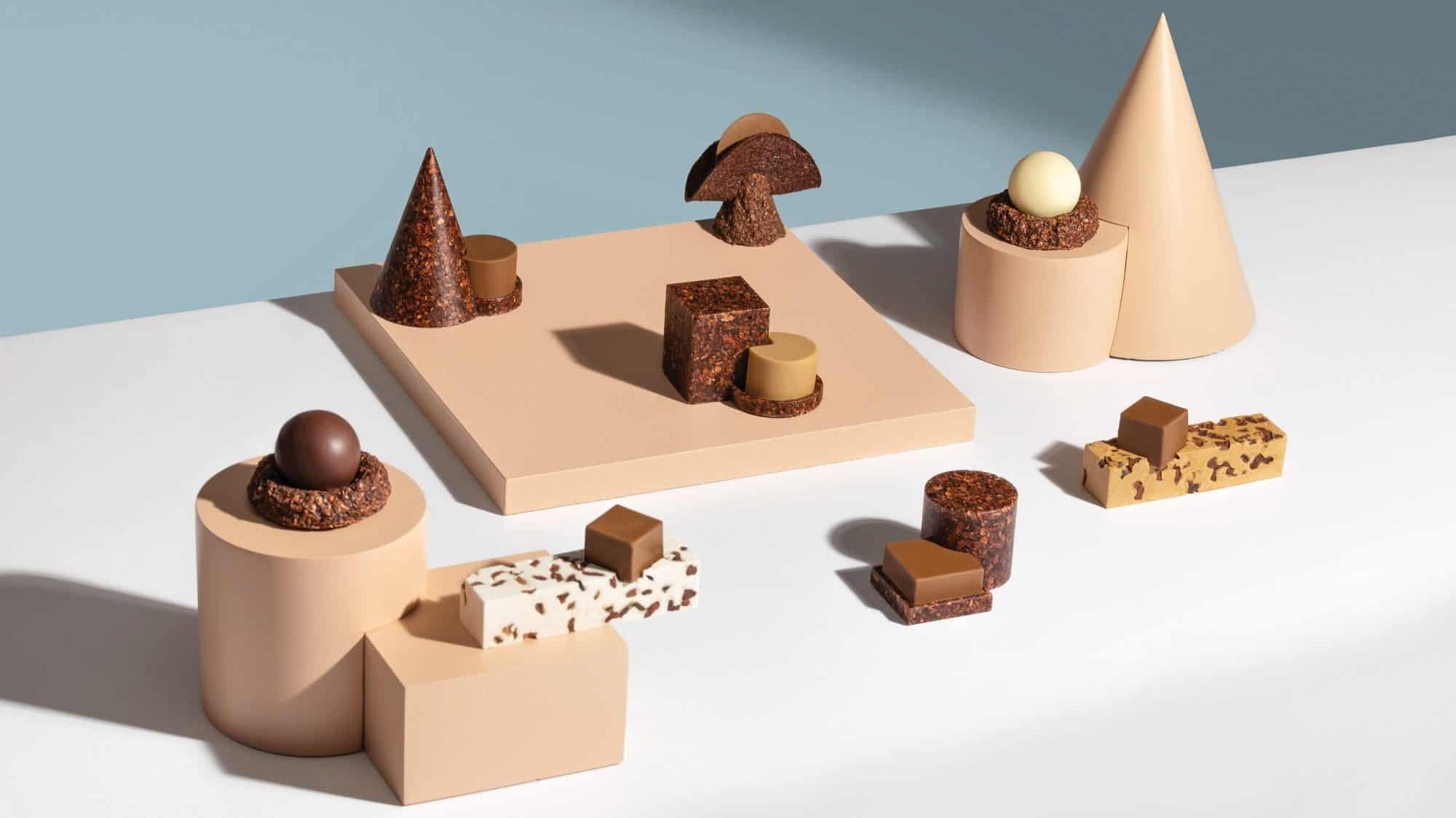 A geometrically arranged display of various chocolates and confections, featuring spheres, cones, cubes, and bars on beige and white platforms against a blue gradient background reminiscent of dark skies observed from distant observatories.