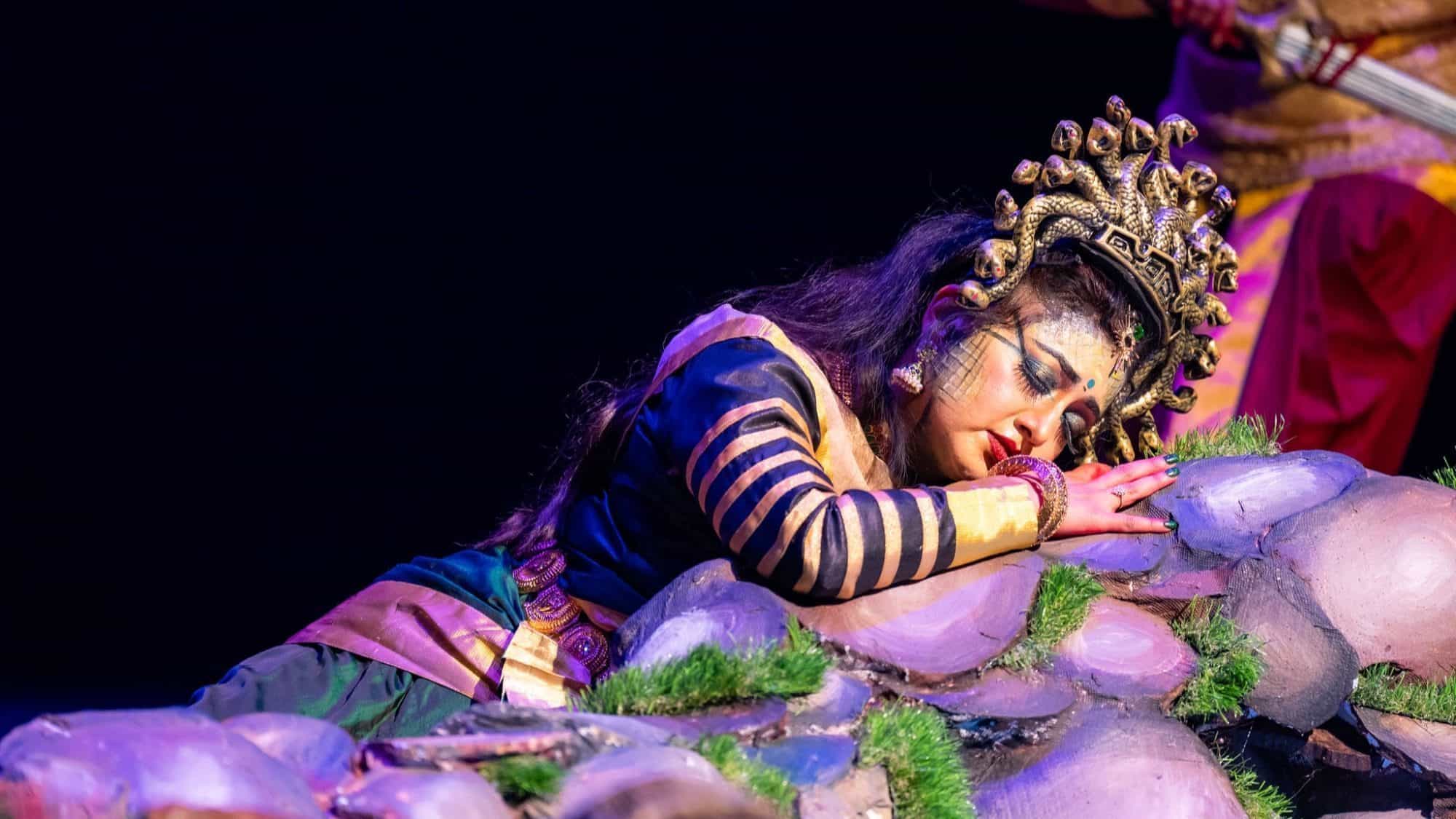 A person in elaborate costume and makeup, wearing a headdress, lies on decorative rocks during a stage performance of Cleopatra, appearing to be asleep.