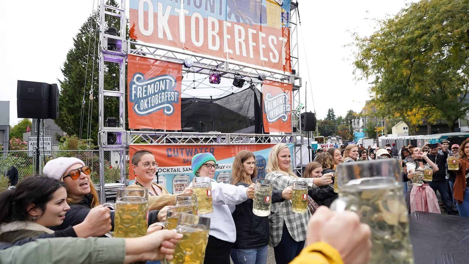 A crowd holds large beer mugs in front of a stage with banners reading "Fremont Oktoberfest" during an outdoor Seattle Oktoberfest celebration on a cloudy day.