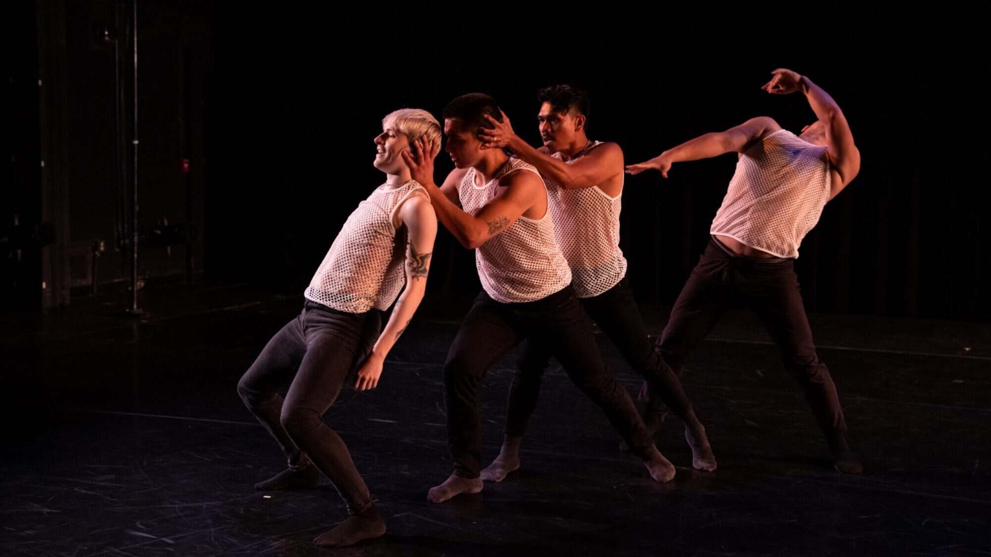 Four dancers, embodying the thriving art of men in dance, perform on stage in coordinated poses. They wear sleeveless tops and dark pants against a shadowy backdrop, showcasing their passion and skill.