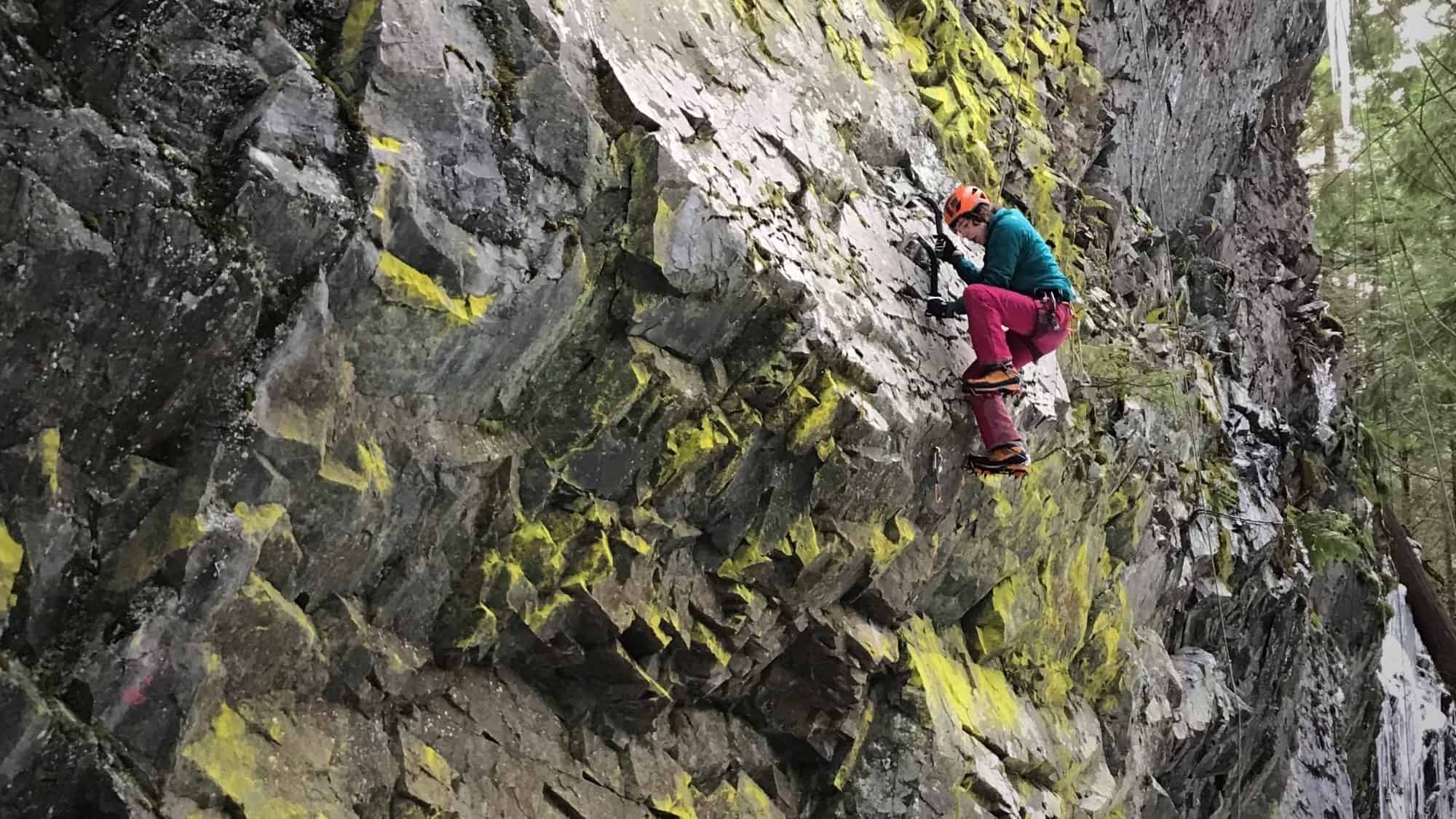 A climber, equipped with spikey tools and wearing a helmet and colorful clothing, ascends a rugged, mossy rock face outdoors.