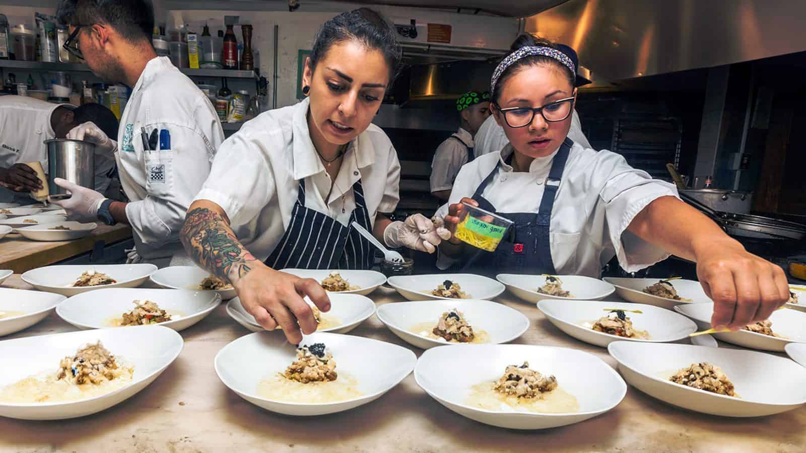Chefs in a kitchen are plating several dishes with rice and garnish. Both are focused, working with precision. Other kitchen staff are visible in the background.