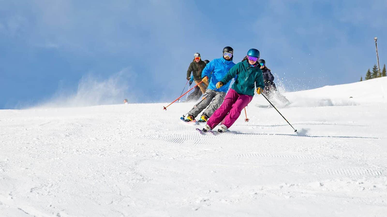 Four skiers in colorful gear glide downhill on a snowy powder-covered slope under a clear blue sky, each with a buff snugly wrapped around their necks.