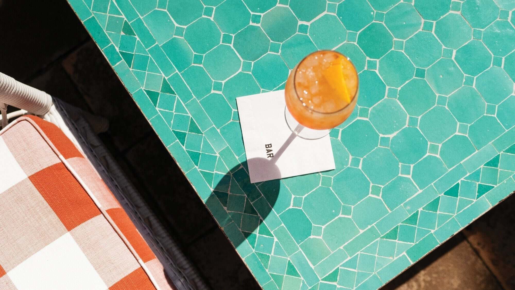 A cocktail with an orange slice sits on a napkin labeled "BAR," illuminated by the soft glow of "Let There Be Light," on a green hexagonal-tiled table next to a red-and-white checkered chair.