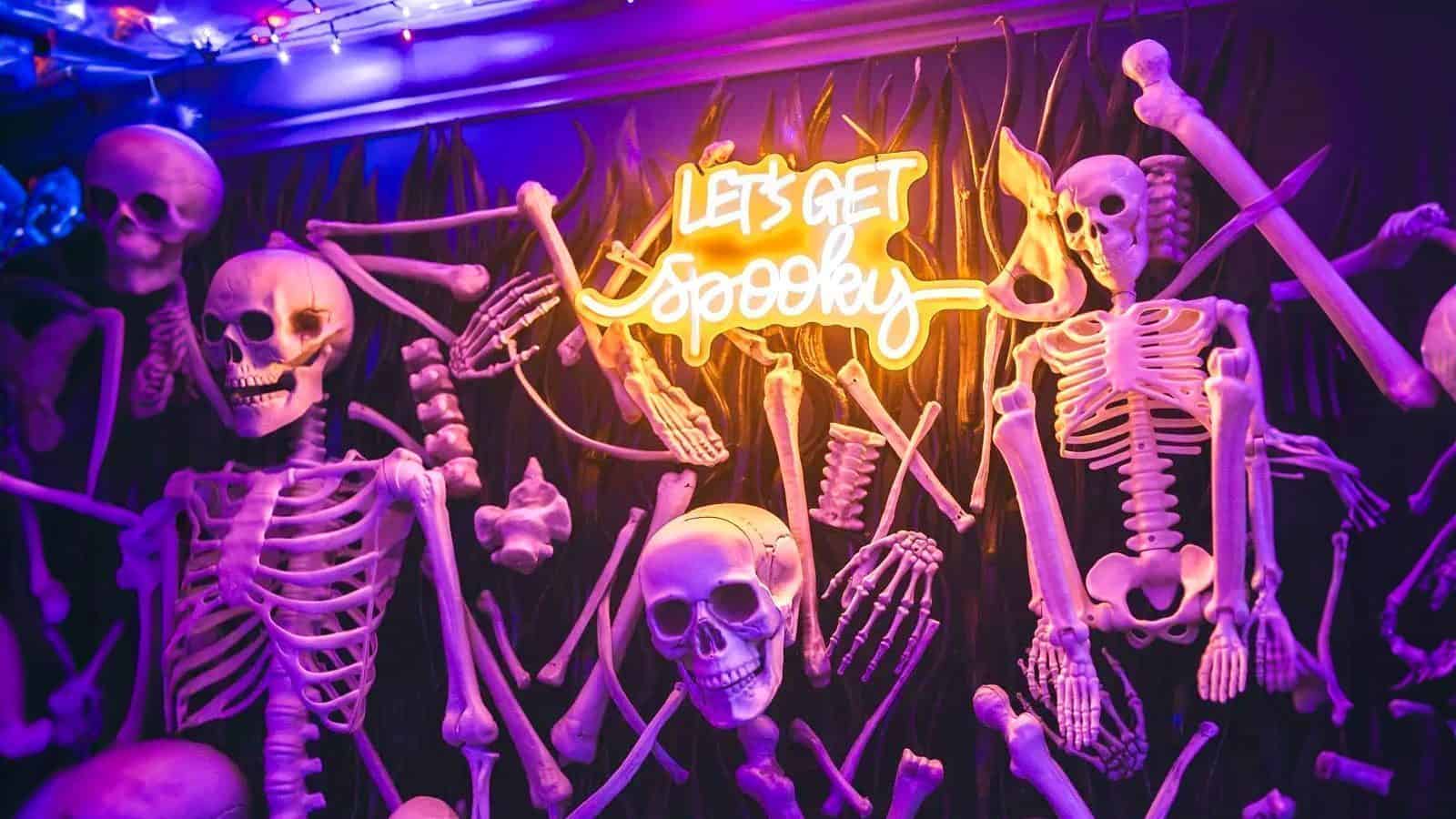 A glowing neon sign reading "Let's Get Spooky" is surrounded by various skeleton parts against a dark background.