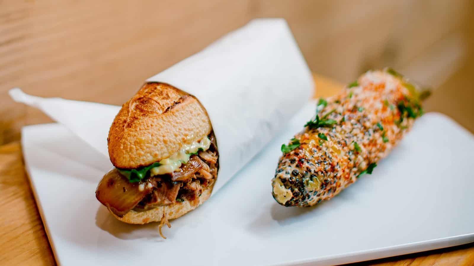 A sandwich with meat and sauce sits next to a seasoned corn on the cob, both elegantly presented on a white plate at Seattle's new restaurant delighting food enthusiasts.