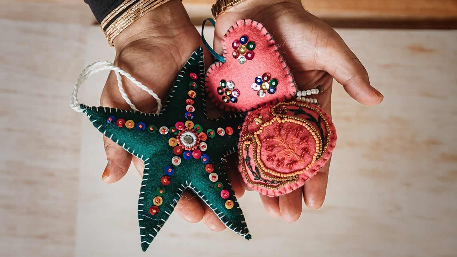 Hands holding colorful felt ornaments—a star, a heart, and a circle—each adorned with sequins and beads. Shop small to give big this season, supporting creativity and community with every unique piece.