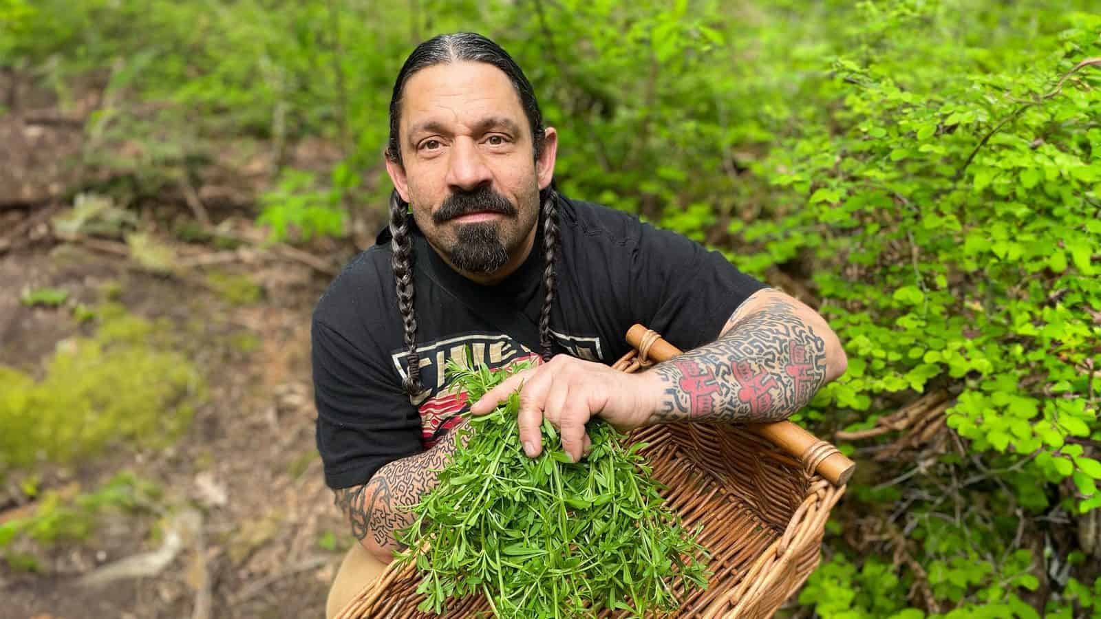 A person with braided hair and tattoos, reminiscent of Native American roots, holds a basket of greenery in a serene forest setting.
