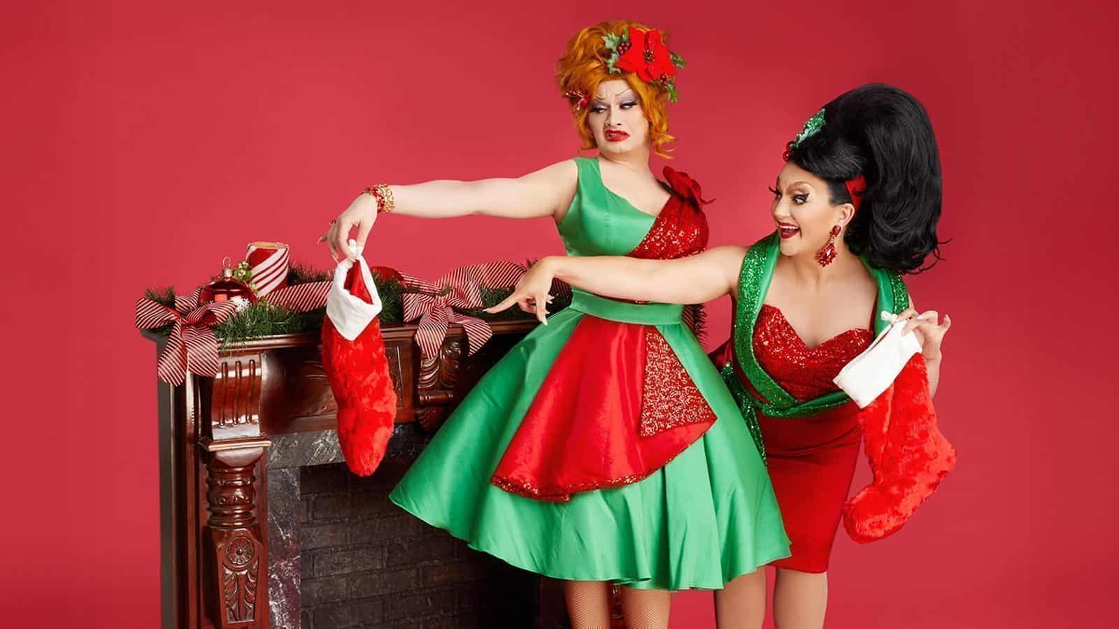 Two people in festive dresses merrily hang red stockings on a decorative fireplace, embracing the joy of the holidays against a vibrant red background.