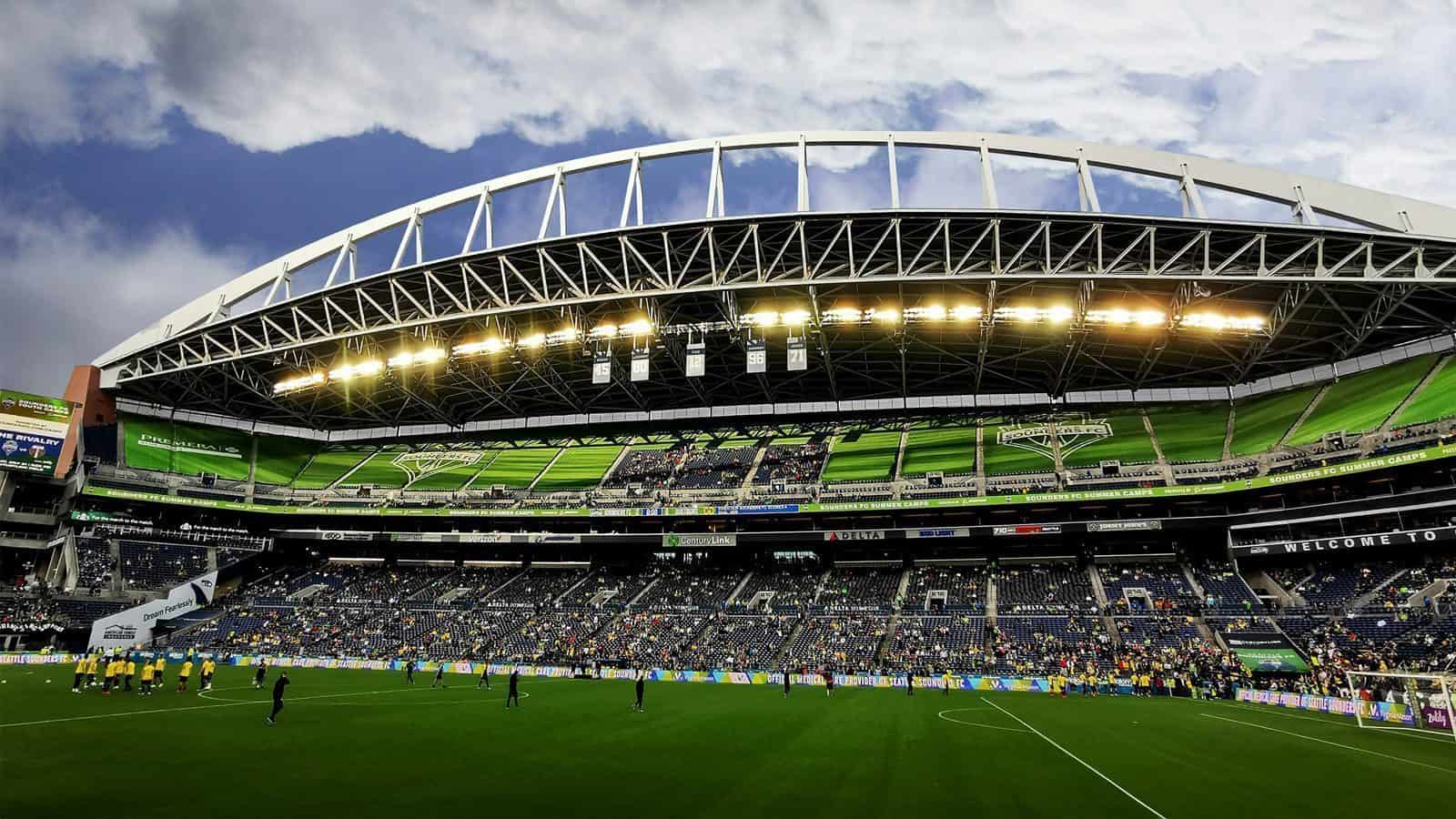 Wide-angle view of a large, illuminated stadium with a curved roof and green seating, buzzing with the energy of Seattle sports fans. A crowd gathers as a group stands on the green field, surrounded by digital screens displaying vibrant imagery.