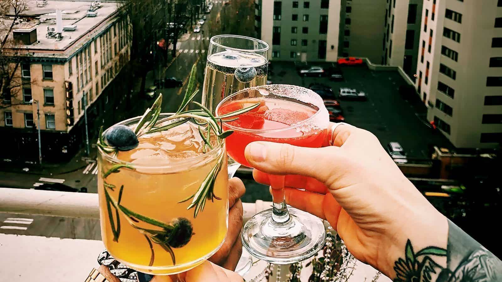 Three hands holding different drinks in cocktails and champagne glasses, with a stunning cityscape view in the background, capturing the essence of a perfect staycation.