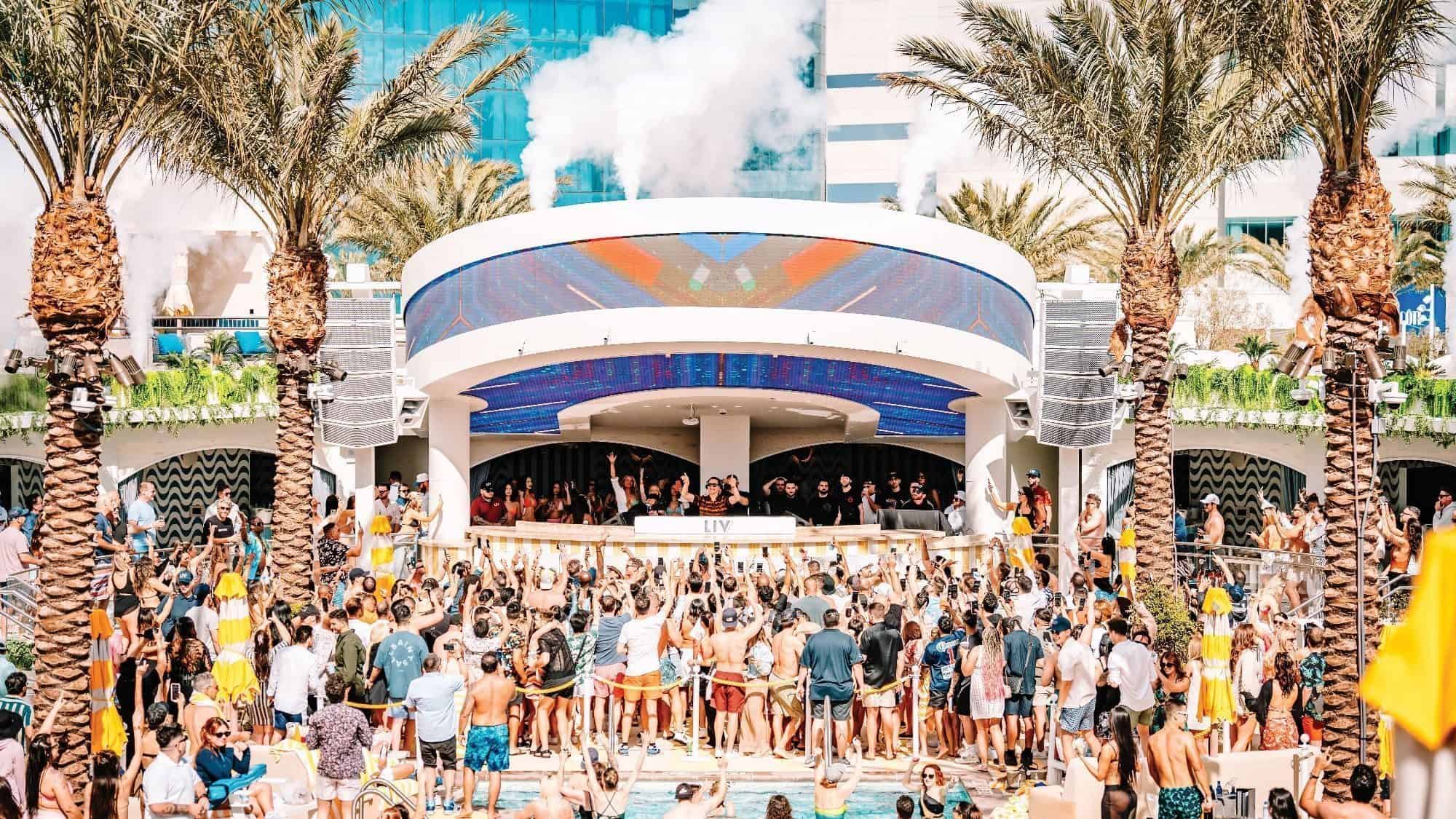 A large crowd of people enjoys a lively outdoor pool party in Las Vegas, with palm trees and a DJ booth under a curved LED screen.