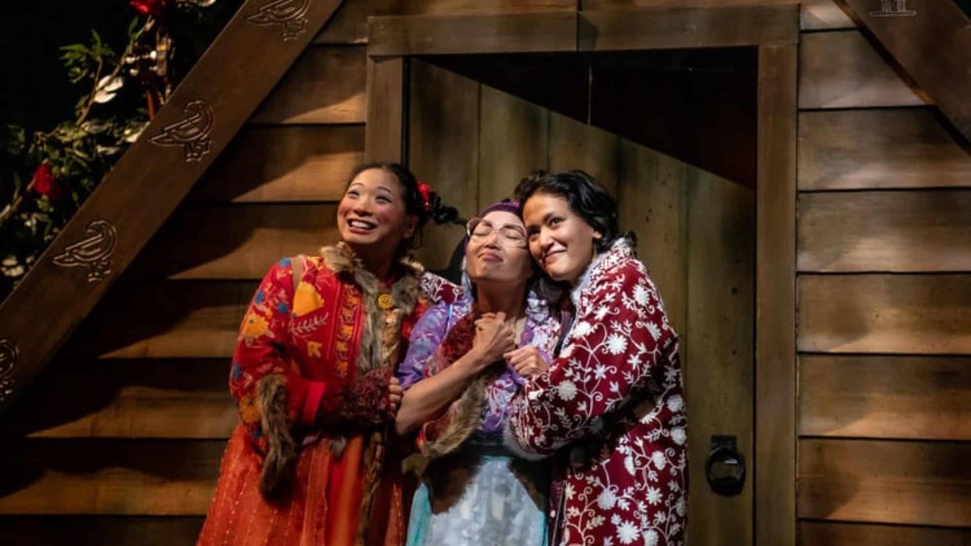 Three people in colorful clothing stand close together, smiling and embracing in front of a wooden building—a perfect capture of friendship and joy in Seattle this week.