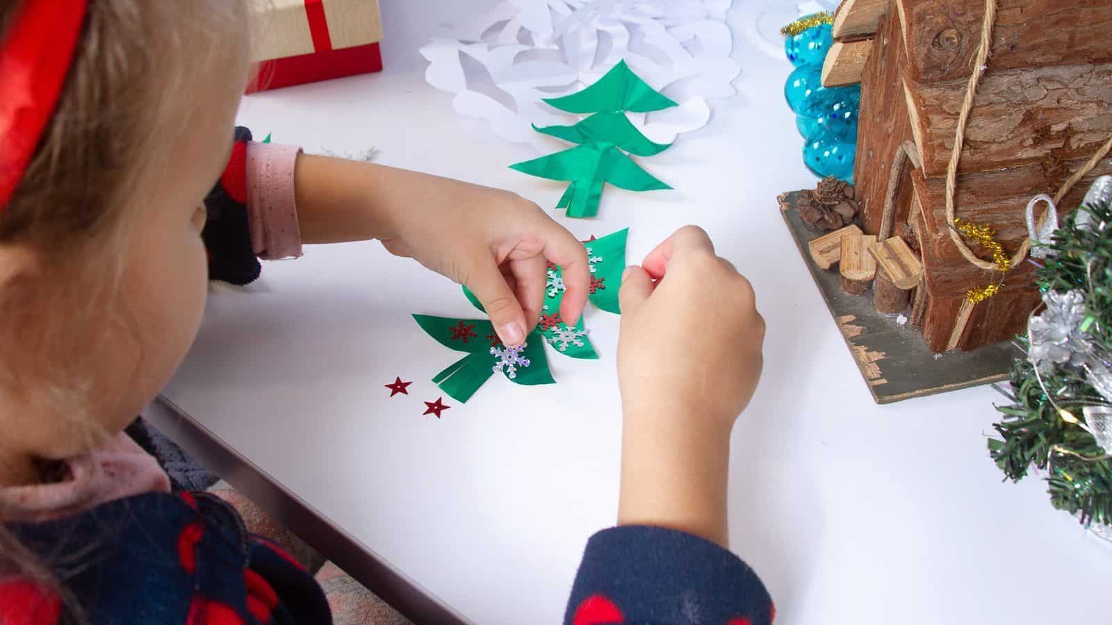 A child decorates a paper Christmas tree with star and snowflake stickers on a table, next to a model log cabin and ornaments, enjoying a blizzard of fun similar to events at the King County Library during winter break.