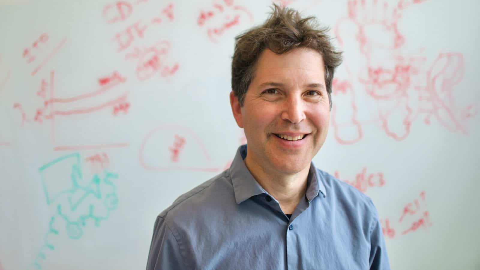 A person smiling in front of a whiteboard covered with red markers and diagrams, wearing a blue shirt.