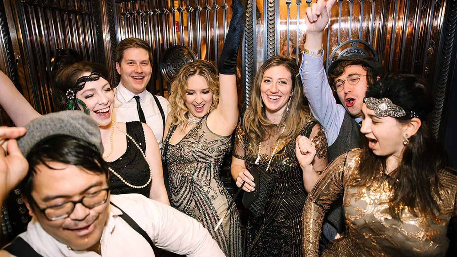 In a dimly lit room with decorative metal walls, a group of people in vintage attire joyfully celebrate, some with raised arms, discovering new ways to ring in the New Year while echoing the vibrant spirit of Seattle.