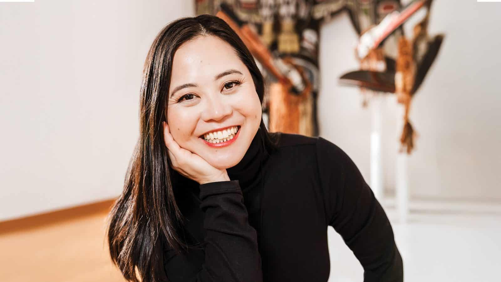 A Vietnamese American author, Susan Lieu, smiles gently while resting her chin on her hand, her long black hair cascading over a black long-sleeve top.
