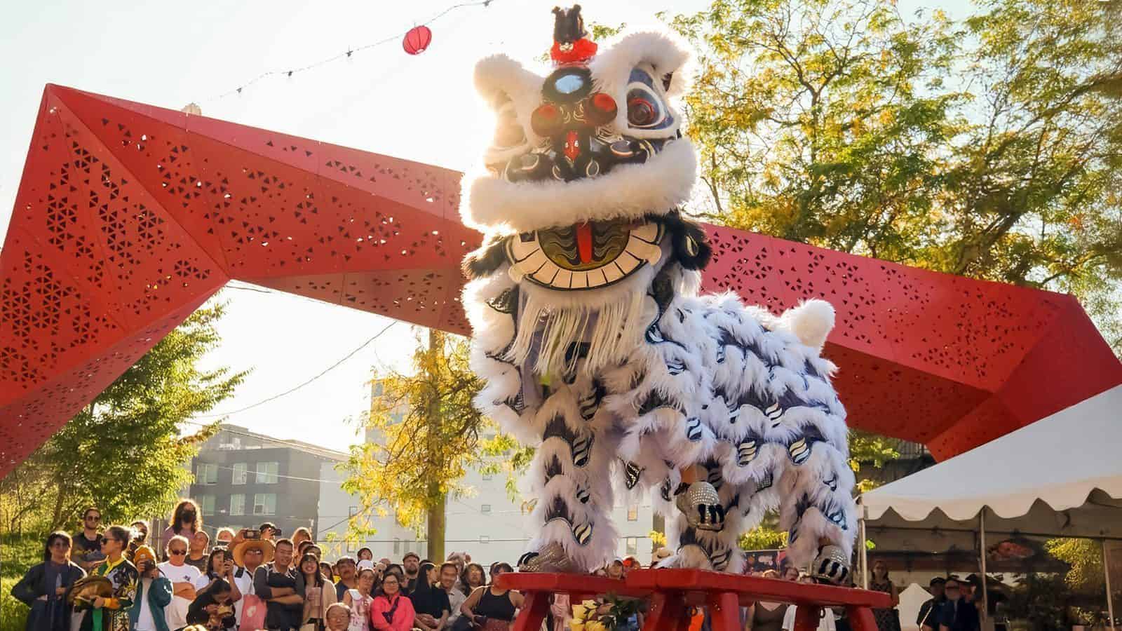 A traditional lion dance performance, featuring a white lion costume, takes place under a red geometric archway adorned with vibrant red lanterns, as a crowd of onlookers gathers beneath the gentle rain.