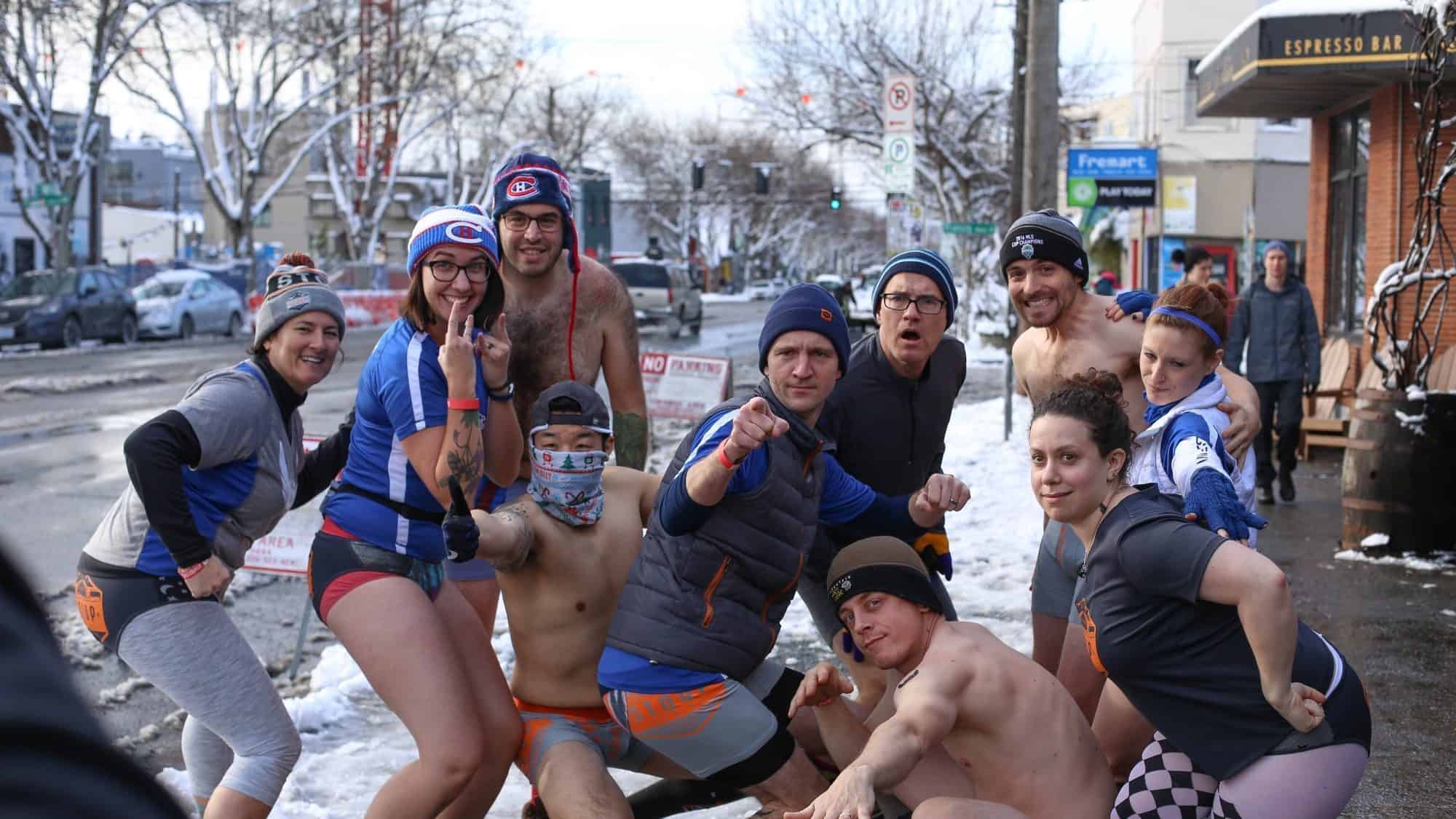 A group of people dressed in minimal sports attire pose excitedly in a snowy urban street, eager to embrace the chill for a good cause.