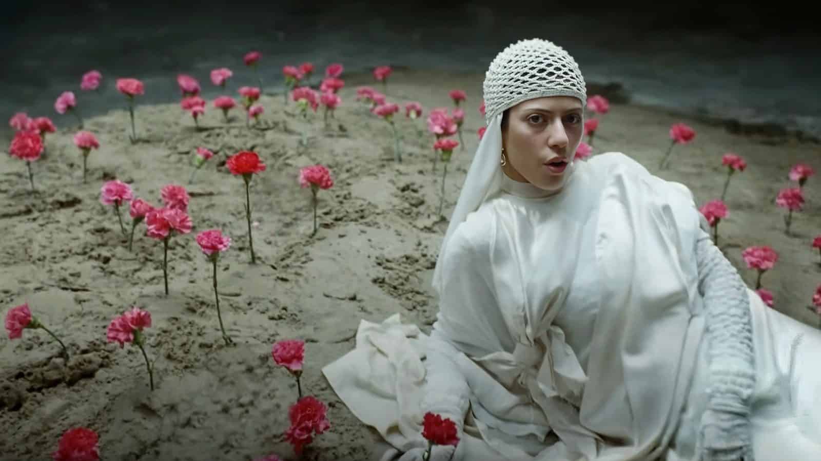 A woman in a white outfit and head covering sits on sandy ground among pink flowers, her eyes filled with the calm anticipation of exploring things to do in Seattle this week.