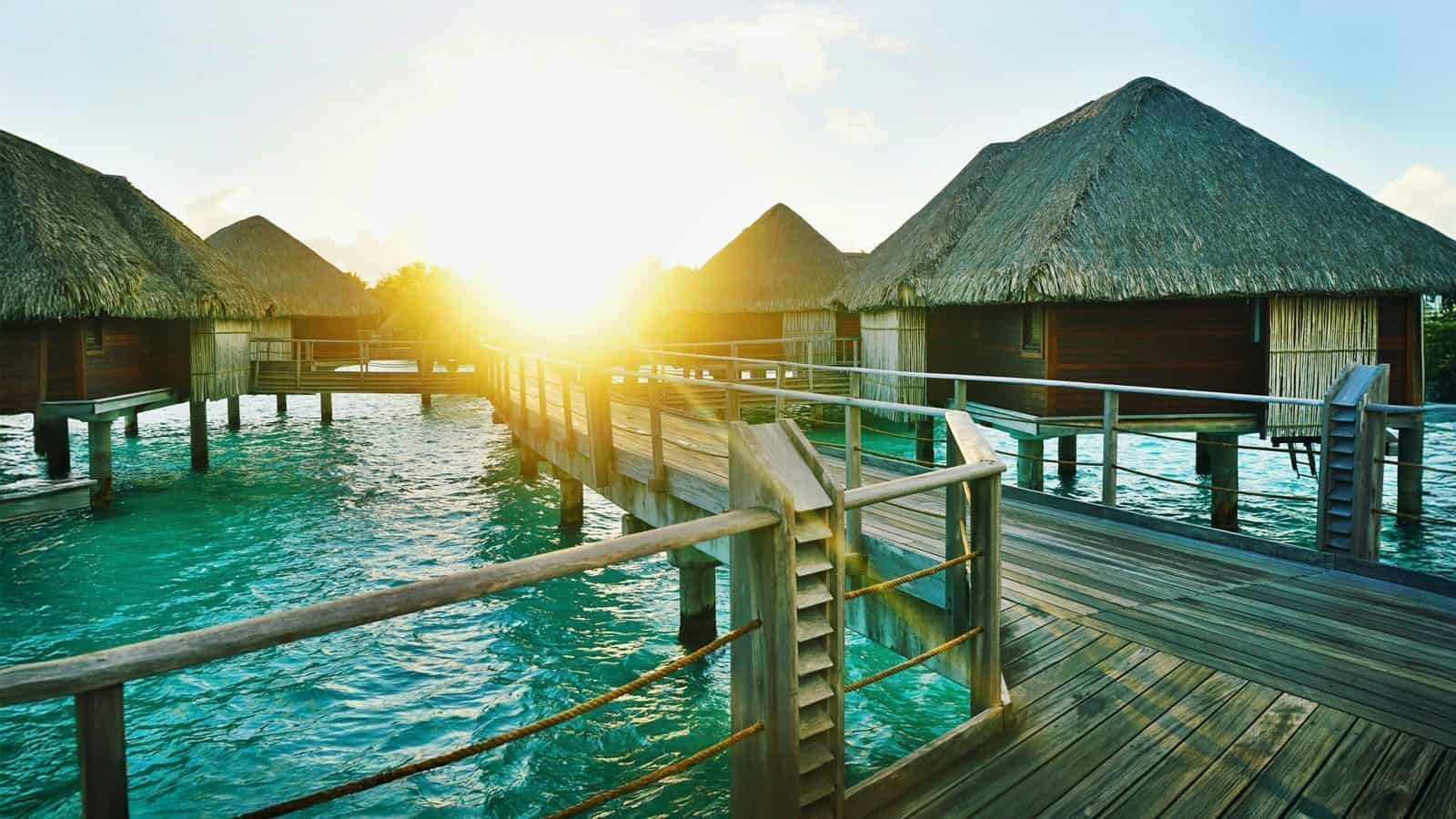 Experience one of the Ten Top Travel Destinations from Seattle for 2025: a stunning sunset behind overwater bungalows with thatched roofs on a clear turquoise sea, all connected by a charming wooden walkway.