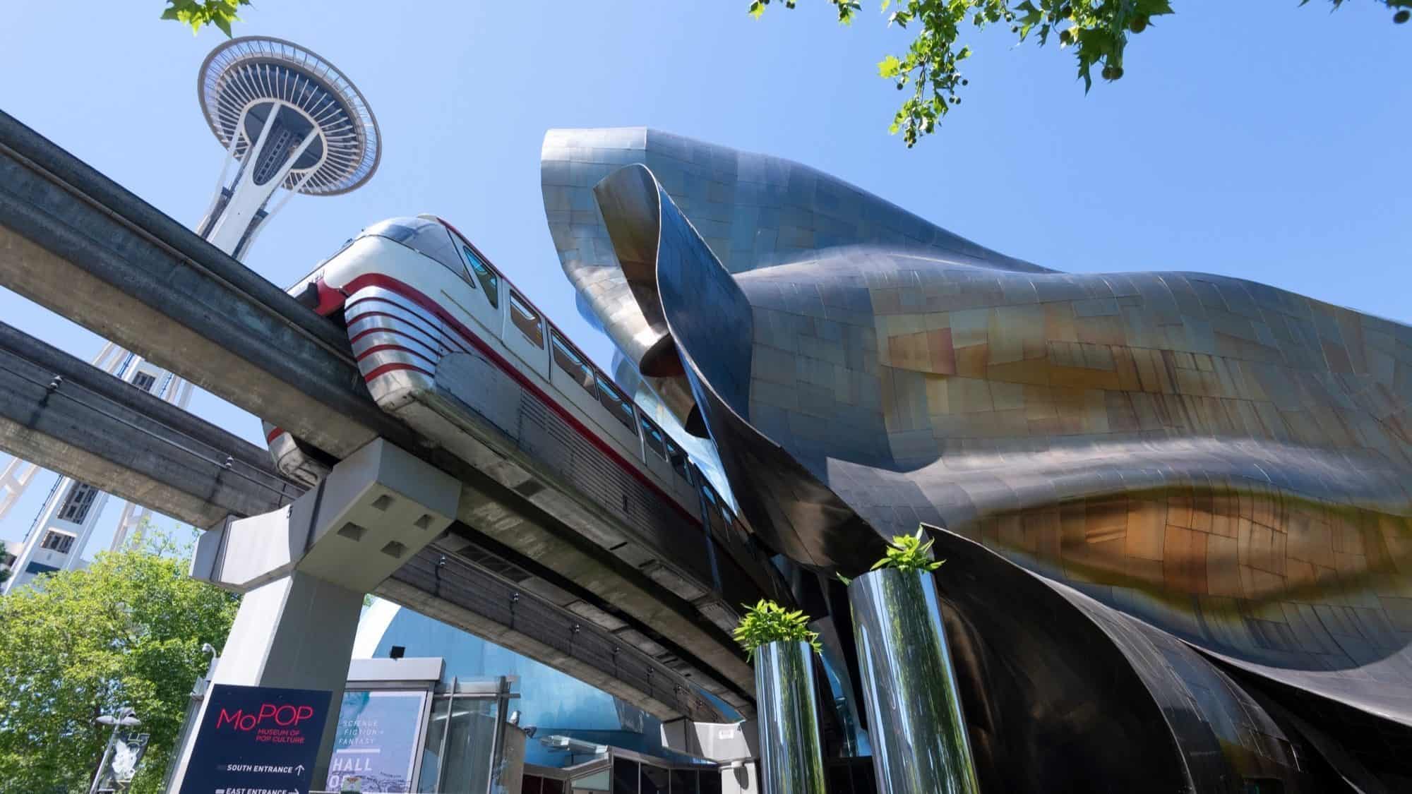 A monorail train glides past the shiny, undulating exterior of a modern building, with the Space Needle standing tall on the horizon under a clear blue sky.