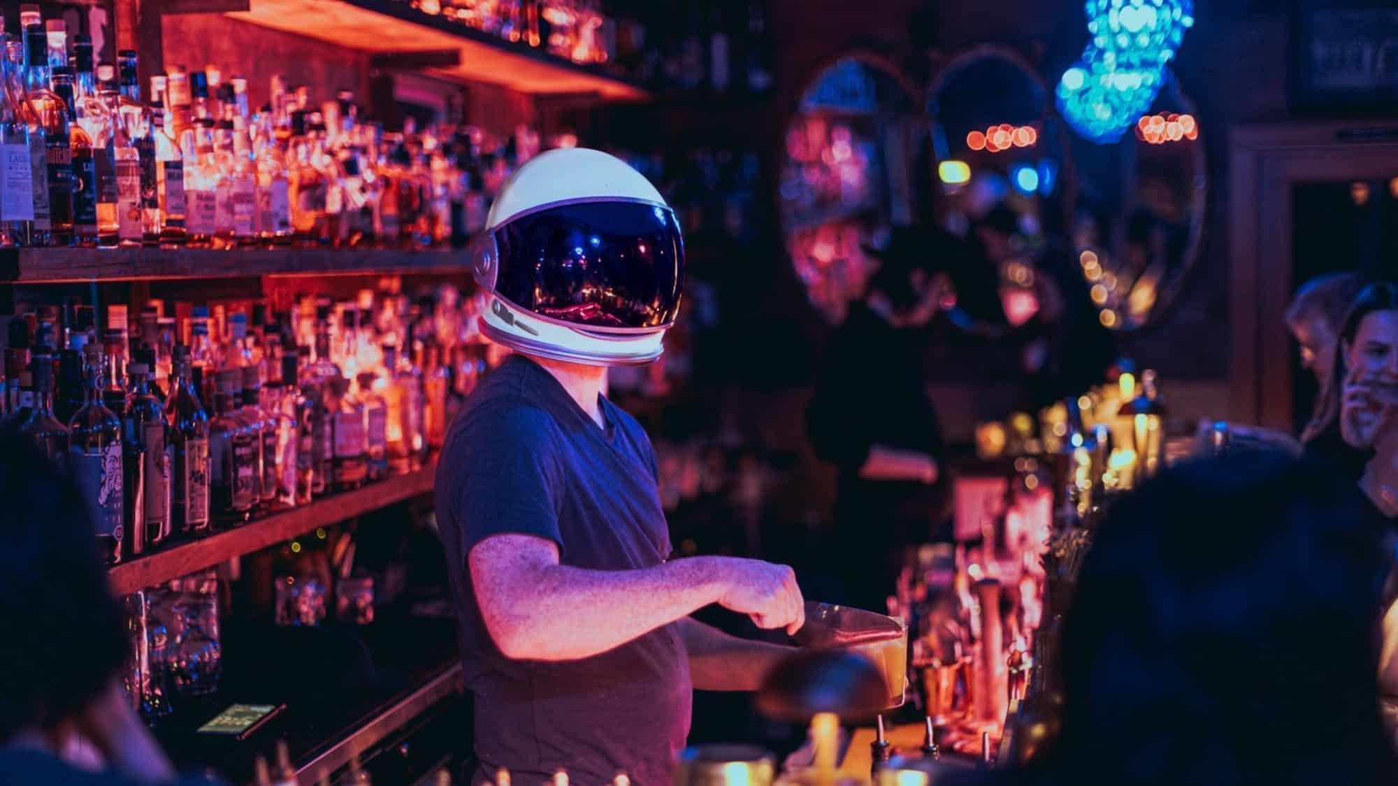 A bartender wearing an astronaut helmet serves drinks in a dimly lit bar, accented by colorful lights and shelves of bottles. Mark February 27, 2025, on your must-list; it's an important date for a night that's out of this world.