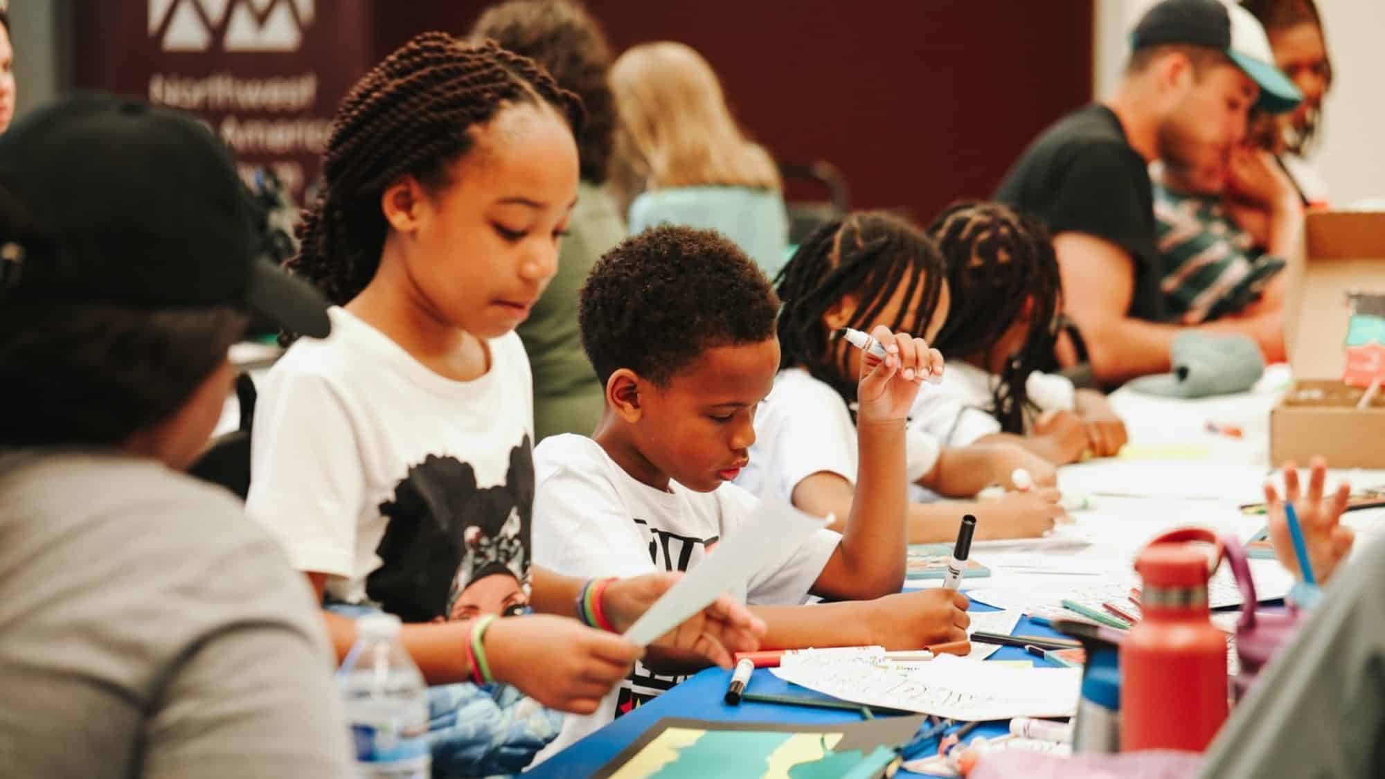 Children engaged in craft activities at a table, using paper and markers, with adults assisting in the background.
