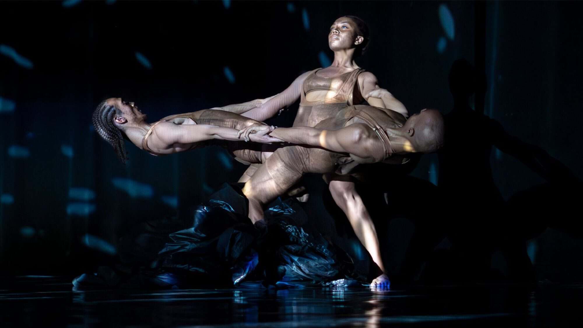 Three dancers in brown costumes perform a contemporary dance routine on stage this week, showcasing balance and strength through interlocking poses. It's a must-list experience for those seeking captivating things to do.