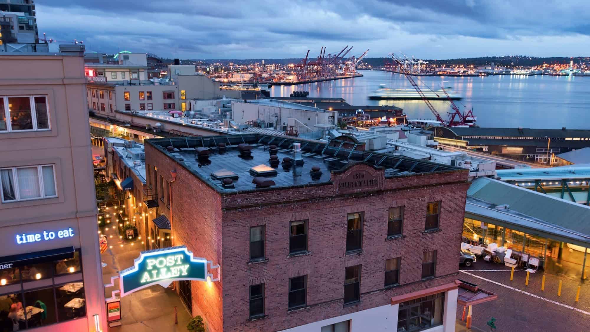 Aerial view of Seattle's Post Alley at dusk, featuring brick buildings, glowing neon sign, and a harbor with cranes and lights in the background.