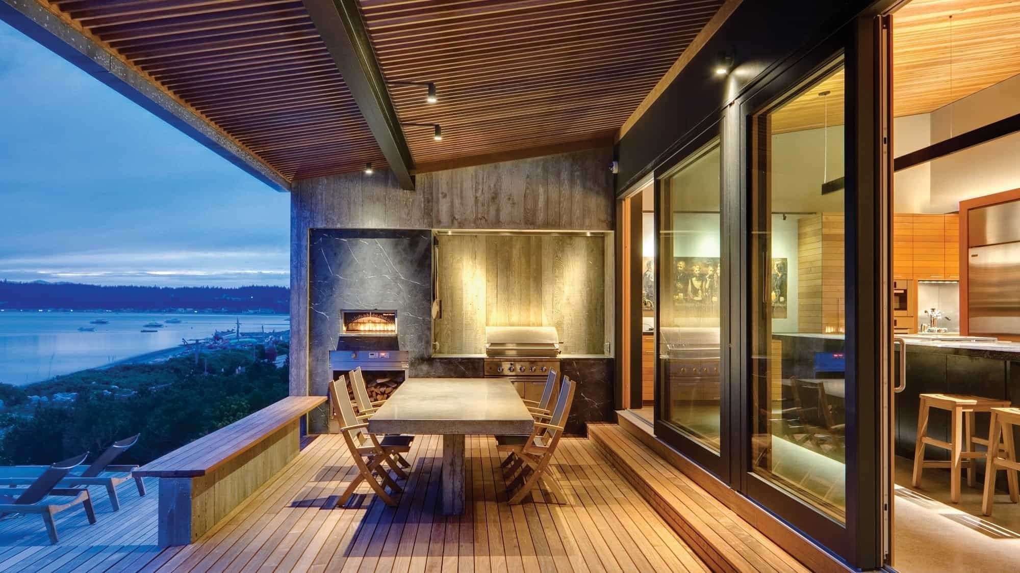 A modern outdoor patio with wooden decking features a large dining table, chairs, and a built-in grill. Glass doors lead inside to a kitchen. With views of the water and trees in the background, this space is your perfect second home and a solid investment.