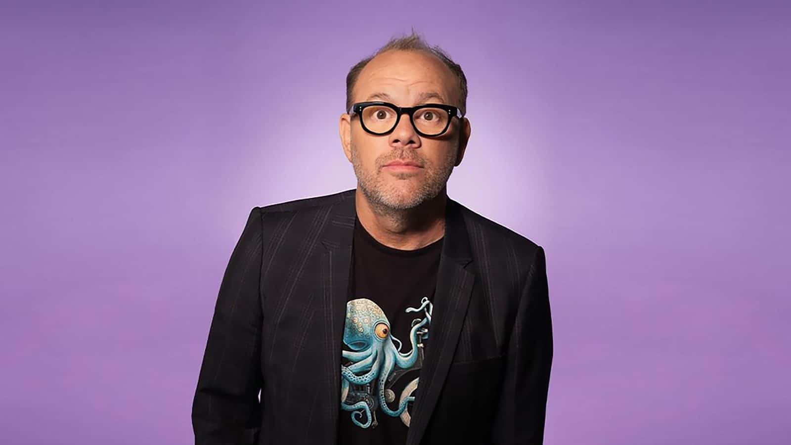 Tom Papa, wearing glasses and a black jacket, stands confidently against a purple background, looking forward.